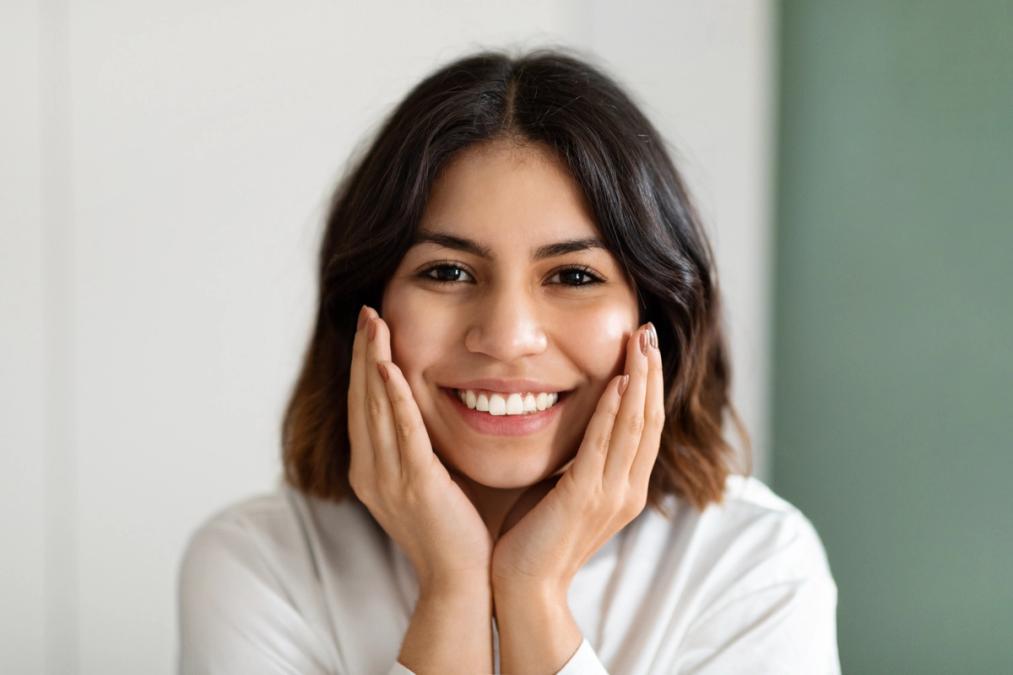 A young woman with dark hair smiling, with her hands cupping her face.
