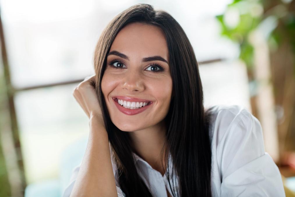A smiling woman with long dark hair, wearing a white shirt, resting her chin on her hand.