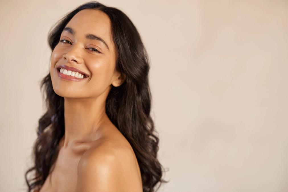 Smiling woman with long dark wavy hair, clear skin, and bare shoulders.
